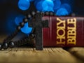 Catholic cross on a rosary and a book - the Holy Bible on a wooden table. Dark blue background with twinkling lights. Faith, Royalty Free Stock Photo