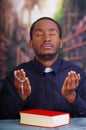 Catholic priest wearing traditional clerical collar shirt sitting with folded hands holding rosary while praying, book Royalty Free Stock Photo