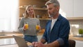 Caucasian adult woman pour orange juice give glass husband man working online with laptop in home kitchen clinking Royalty Free Stock Photo