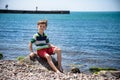 Caucasian baby boy is sitting at the beach. Cute child is playing with roks and shellfishes muscle-fish on the sand in summer day