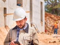 Caucasian bearded civil engineer or construction worker in white hardhat uses his pnone opposite construction site Royalty Free Stock Photo