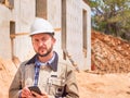 Caucasian bearded civil engineer or construction worker in white hardhat uses his pnone opposite construction site