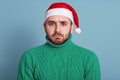 Caucasian bearded man wearing stylish green sweater and santa hat isolated over blue background, having upset facial expession,