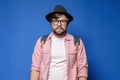 Caucasian bearded, shaggy man in hat, glasses and casual clothes looks calmly into the camera. Blue background.