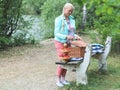A Caucasian blonde woman stands near a bench in a forest with a lake and, having opened a picnic basket Royalty Free Stock Photo