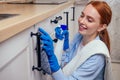 Caucasian beautiful red-headed female in casual clothes wearing rubber glove and holding chemica spray in kitchen