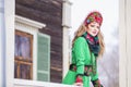 Caucasian Blond Girl in Fashionable Green Dress and Kokoshnik with Flowery Pattern and Beads. Posing Against Old wooden House in Royalty Free Stock Photo