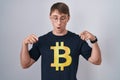 Caucasian blond man wearing bitcoin t shirt pointing down with fingers showing advertisement, surprised face and open mouth