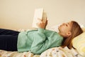 Caucasian blond teenage girl is lying on the bed and reading book