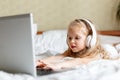 Caucasian blonde child girl use laptop in headphones for doing homework watching videos or cartoons. Girl sitting on bed
