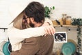 Caucasian blonde woman in white hat hugging her partner with dark hair and beard holding ultrasound photo. Expecting