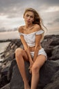Caucasian blonde young woman relaxing on the rocky beach. Sensual girl looking at the camera, sitting on the rocks Royalty Free Stock Photo
