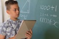 Caucasian boy standing at chalkboard in classroom and using tablet during maths lesson Royalty Free Stock Photo