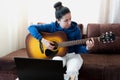 Caucasian brunette woman sitting on sofa and playing acoustic guitar at home. On-line lesson