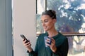 Caucasian businesswoman using smartphone while holding coffee mug standing at workplace Royalty Free Stock Photo