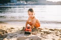 Caucasian child boy playing toy red tractor, excavator on a sandy beach by the river in red shorts at sunset day Royalty Free Stock Photo