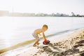 Caucasian child boy plays toy red tractor, excavator on sandy beach by the river in shorts at sunset day Royalty Free Stock Photo