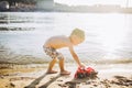 Caucasian child boy plays toy red tractor, excavator on sandy beach by the river in shorts at sunset day Royalty Free Stock Photo
