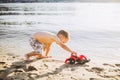 Caucasian child boy plays toy red tractor, excavator on sandy beach by the river in shorts at sunset day Royalty Free Stock Photo