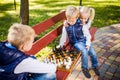 Caucasian children play chess on wooden chessboard in park bench. Brothers and sister big friendly active mental family spend time Royalty Free Stock Photo
