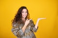 Curly woman pretending to hold something on the palm pointing with a finger isolated on yellow studio background Royalty Free Stock Photo