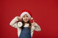 Caucasian smiling little girl in Santa Claus hat, showing alarm clock with midnight on the dial and thumb up at camera Royalty Free Stock Photo