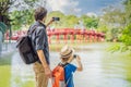 Caucasian Dad and son travelers on background of Red Bridge in public park garden with trees and reflection in the Royalty Free Stock Photo