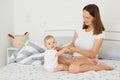 Caucasian dark haired mother and child sitting on white bed, mommy and baby girl playing at home, woman wearing white t shirt and