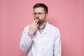 Caucasian doctor in a white coat pensively and anxiously looks to the side, holding hand to face. Pink background.