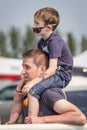 Caucasian father carrying his son on his shoulders outside at an event. Son has sunglasses under his nose