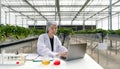 Caucasian female botanical scientist in white gown typing on laptop computer. The table is full of research papers, plant samples