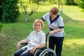 Caucasian female doctor walks with an elderly patient in a wheelchair in the park. Nurse accompanies an old woman on a