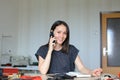 Caucasian female person sitting at leather atelier and making handmade notebook and wallets, talking by smartphone