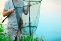 The Caucasian fisherman is standing near the water in the grass and holding the fishing rod and the green landing net with one Royalty Free Stock Photo