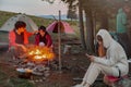 Caucasian friends hikers warming up at fire in camp site in mountains. Evening.