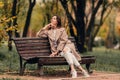 A Caucasian girl in a beige coat and blue jeans sits on a bench in a park in autumn and dreams