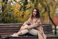 A Caucasian girl in a beige coat and blue jeans sits on a bench in a park in autumn and dreams