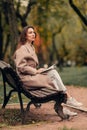 A Caucasian girl in a beige coat and blue jeans sits on a bench in a park in autumn and reads a book Royalty Free Stock Photo