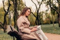 A Caucasian girl in a beige coat and blue jeans sits on a bench in a park in autumn and reads a book Royalty Free Stock Photo