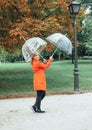 Caucasian girl in red gabardine plays with two transparent umbrellas in a park