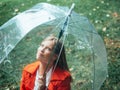 Caucasian girl with red gabardine walks through a park under a transparent umbrella