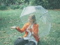 Caucasian girl with red gabardine walks through a park under a transparent umbrella