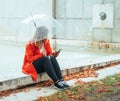 Caucasian girl with red gabardine walks through a park under a transparent umbrella