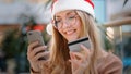 Caucasian girl in santa hat makes internet order using telephone and credit card millennial woman smiling happily buying Royalty Free Stock Photo