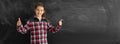 Caucasian girl student or schoolgirl in a shirt stands near a clean chalk board. Raises two index fingers up. Back to Royalty Free Stock Photo