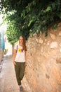 Caucasian girl in white T-shirt with fruits, green pants and gray sneakers is walking along a narrow ancient street with trees and