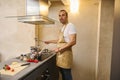 Caucasian handsome young man chef 40s, dressed in beige apron, standing at kitchen counter, cooking delicious dinner for Royalty Free Stock Photo