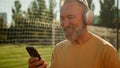 Caucasian happy old man walking stadium football field with headphones listening music smiling texting using phone Royalty Free Stock Photo