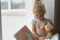 Caucasian kid reads a book, sitting near window and holding bear toy. Royalty Free Stock Photo