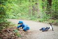 Caucasian little boy toddler in blue jacket, jeans and baseball cap with bike in park playground outside, sitting on ground